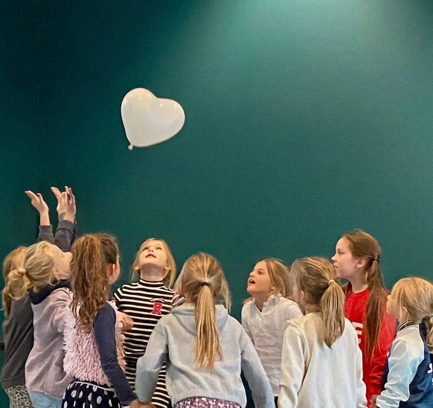 Groep kinderen kijkt omhoog naar een wit hartvormige ballon in een donkergroene zaal