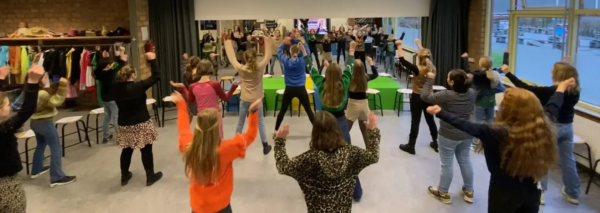 Groep leerlingen met de armen omhoog tijdens een dramaworkshop in de aula