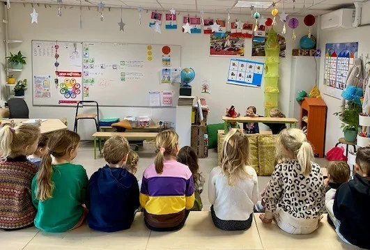 Kinderen zitten op de vloer van het klaslokaal en kijken toe